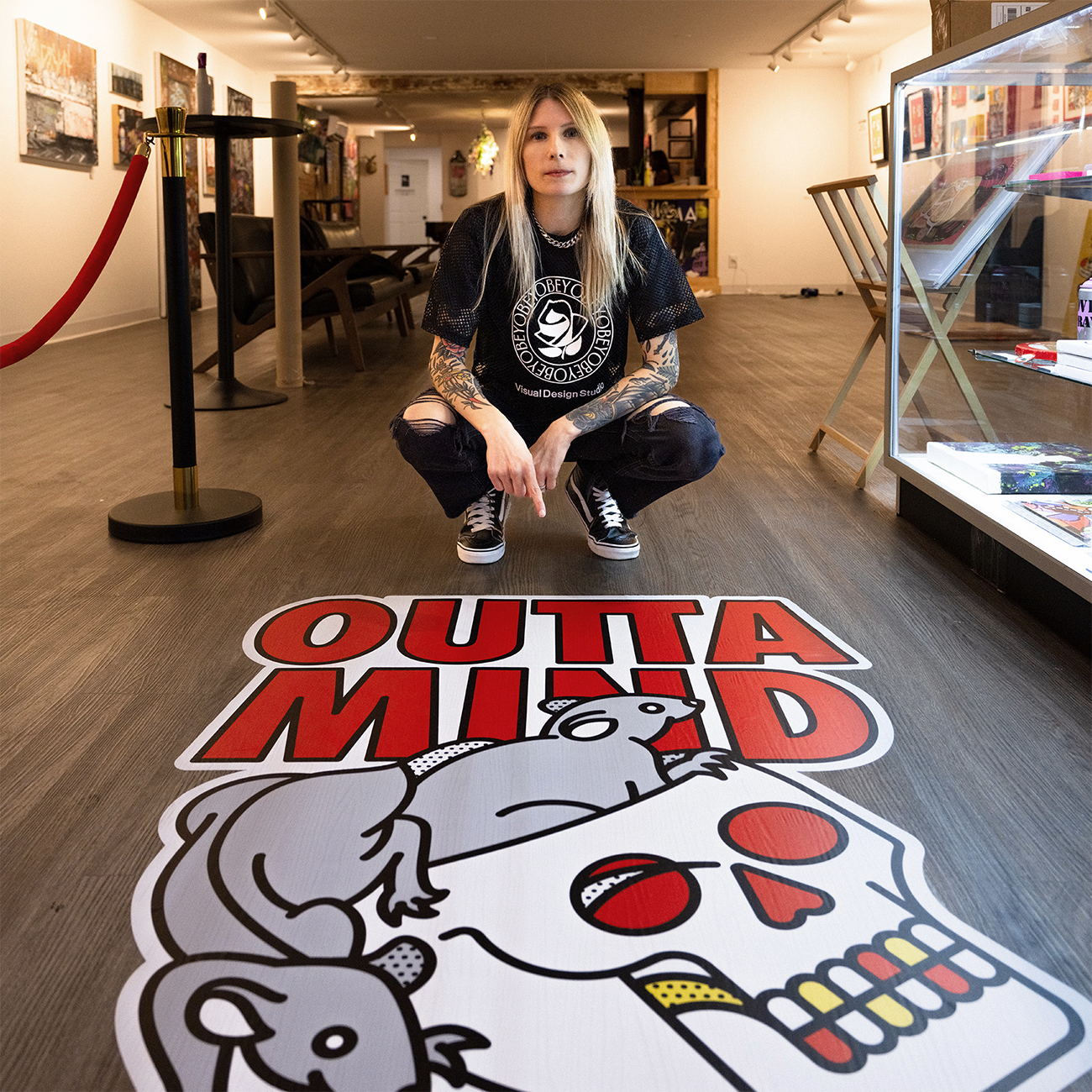 Persona inginocchiata accanto a uno sticker "OUTTA MIND" colorato su un pavimento di una galleria, circondata da opere d'arte.