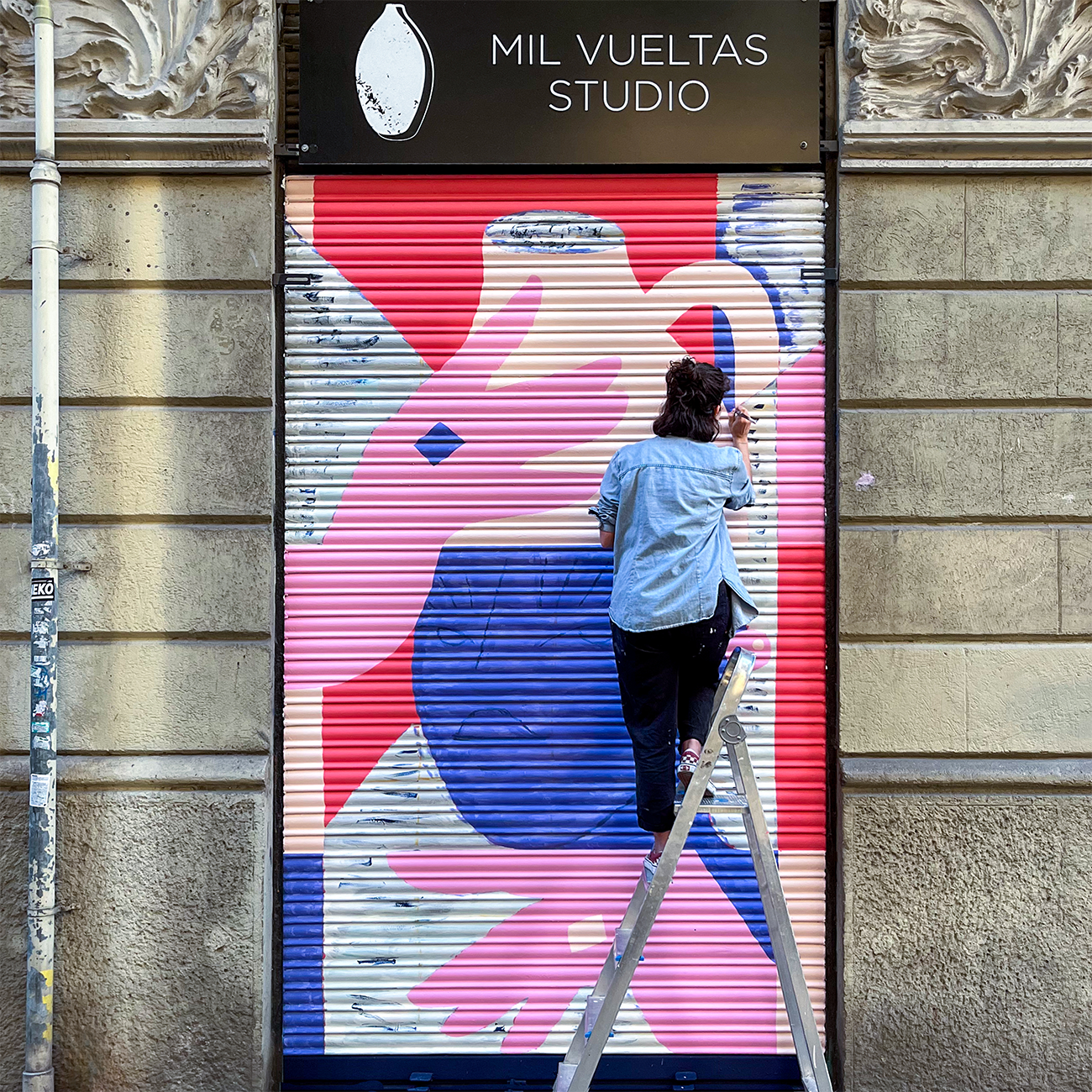Person painting colorful mural on a studio's metal shutter, standing on a ladder against an ornate building facade.