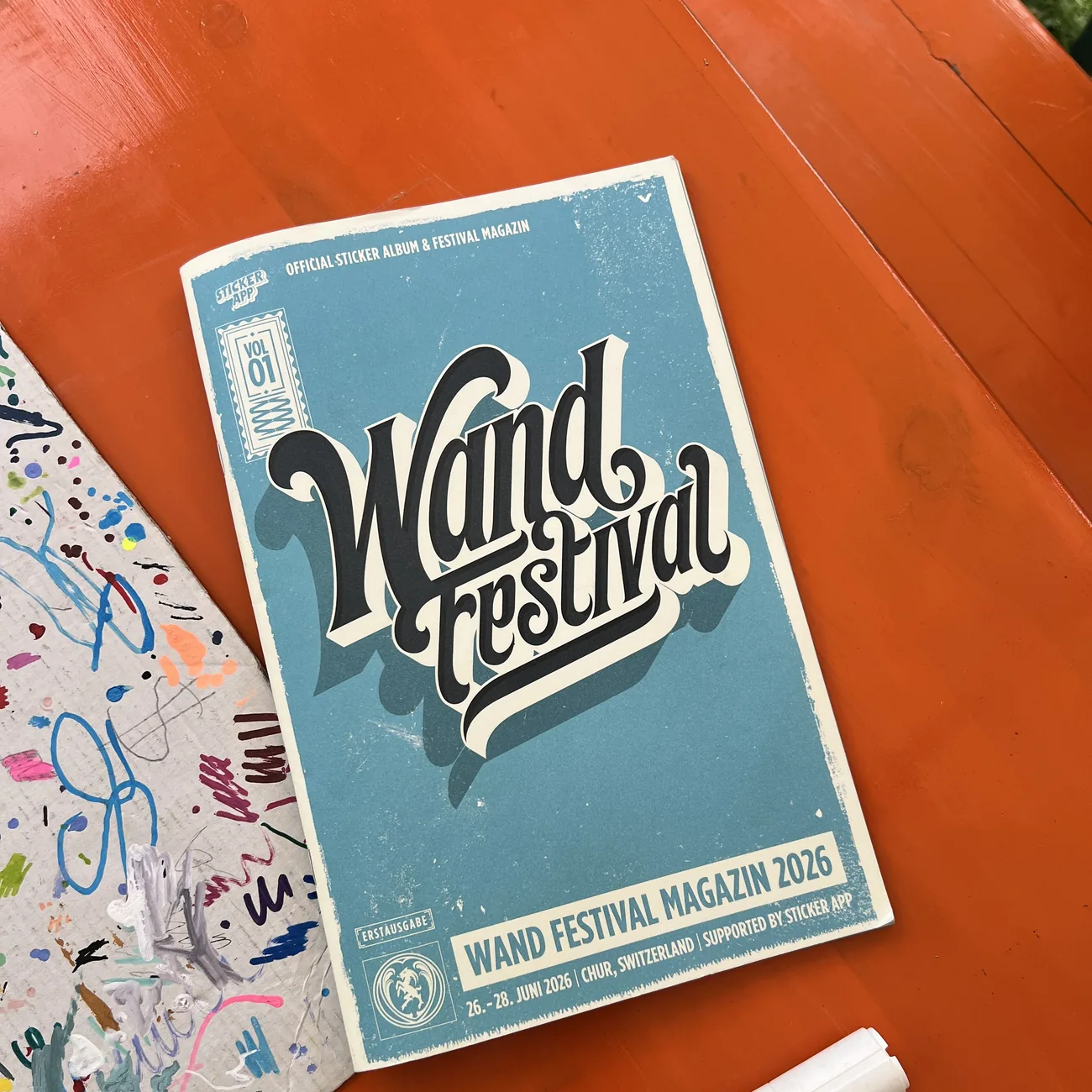 Album ufficiale di adesivi e magazine del Wand Festival 2026, con il supporto di StickerApp, accanto a un marcatore a vernice su un tavolo arancione