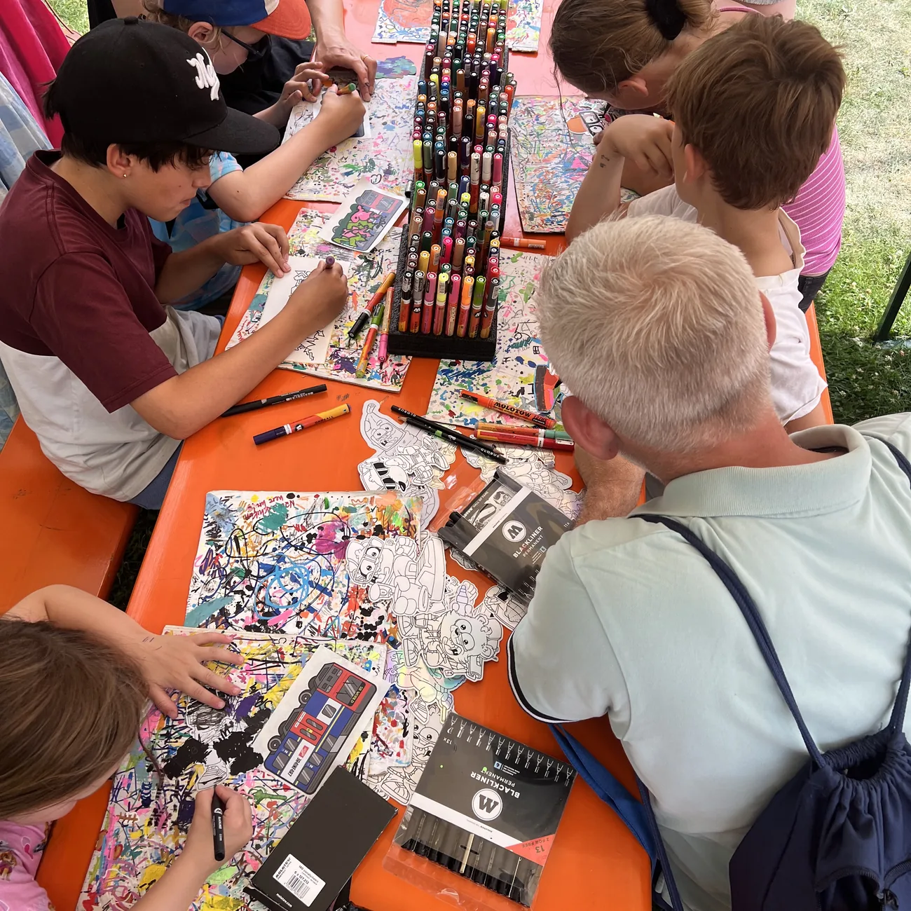 Bambini colorano adesivi sagomati Brainfart attorno a un supporto di marcatori a vernice in un workshop all'aperto durante un festival