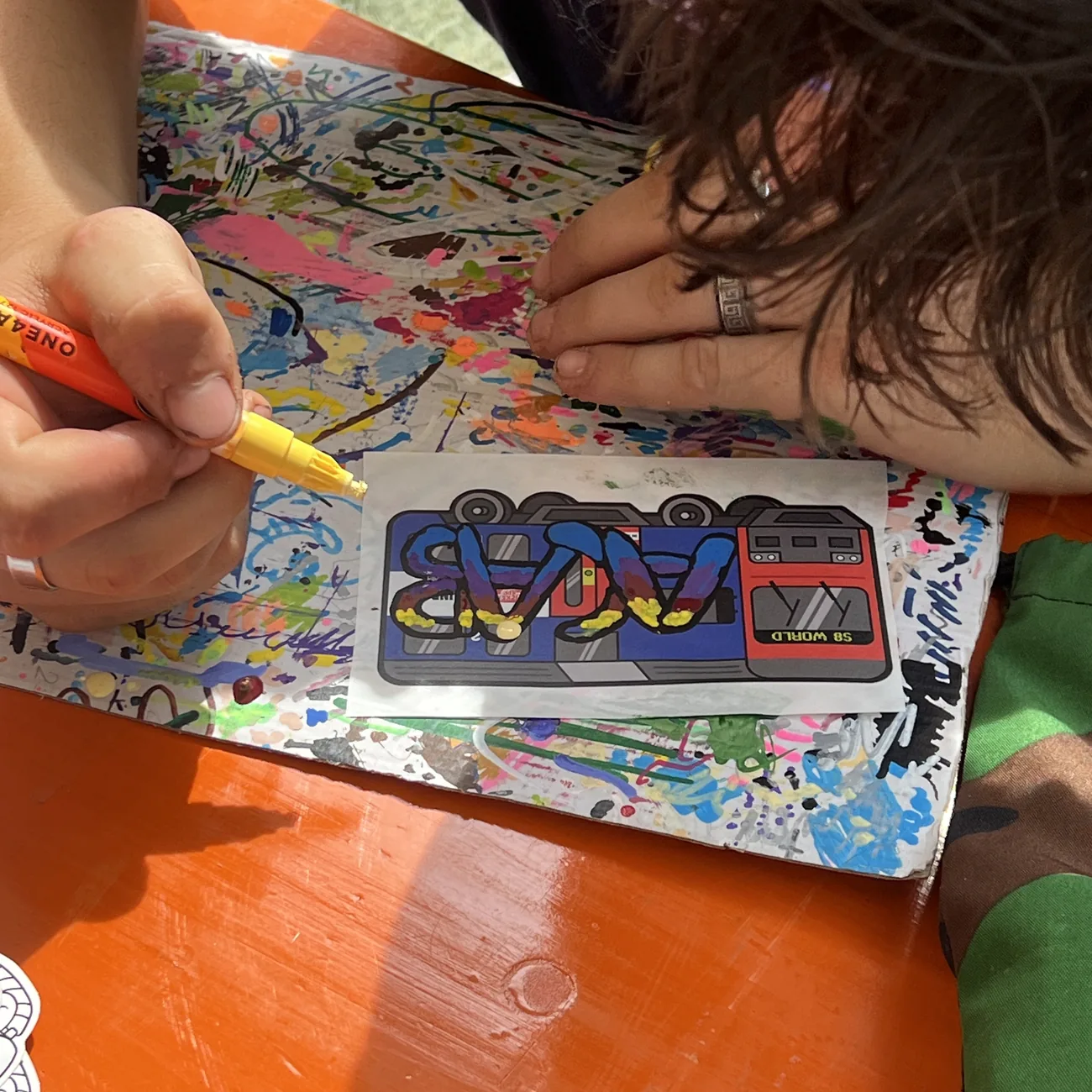 Una mano colora un'etichetta del treno S8 World con un marcatore a vernice giallo al workshop Brainfart x StickerApp