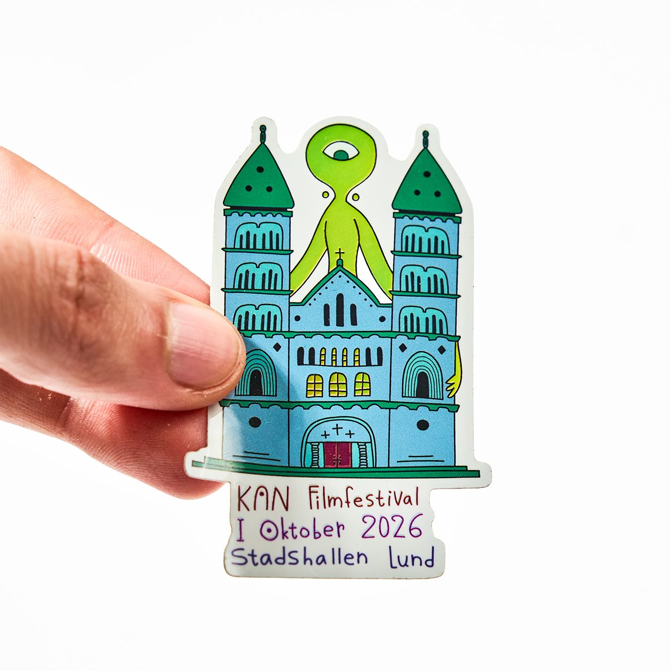 Mano che tiene un adesivo sagomato per il KAN Filmfestival, ottobre 2026 allo Stadshallen di Lund, con una cattedrale e un alieno verde con un occhio solo