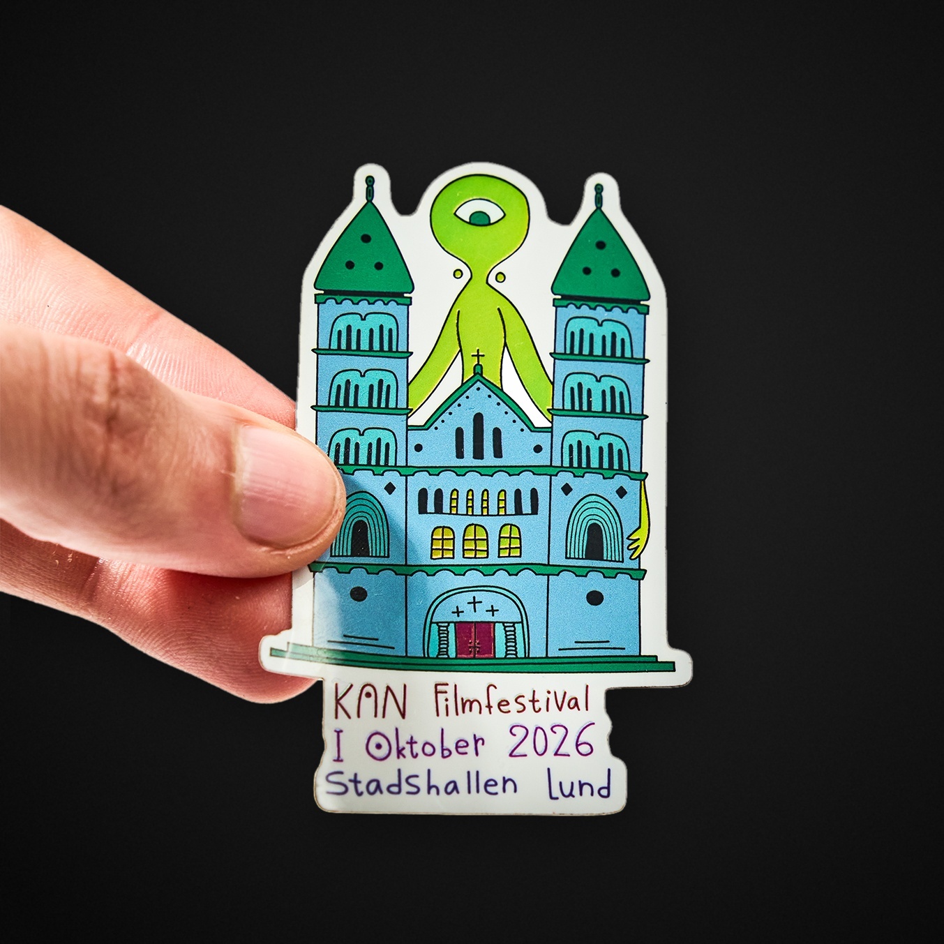 Mano che tiene un adesivo sagomato con una cattedrale e una creatura verde con un occhio solo, con la scritta “KAN Filmfestival I Oktober 2026 Stadshallen Lund”