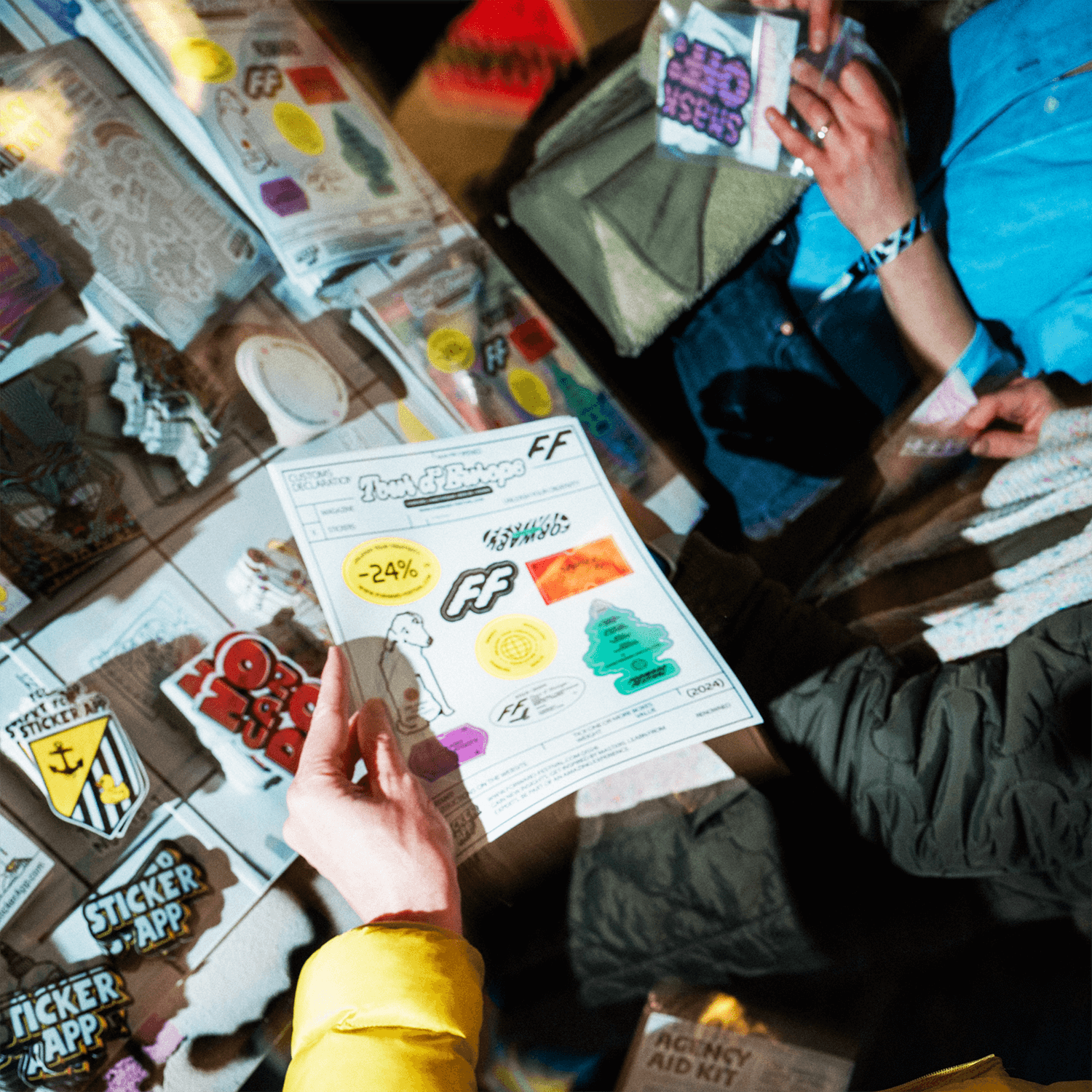 Hands holding a sticker sheet of colorful stickers, surrounded by various sticker designs on a table.