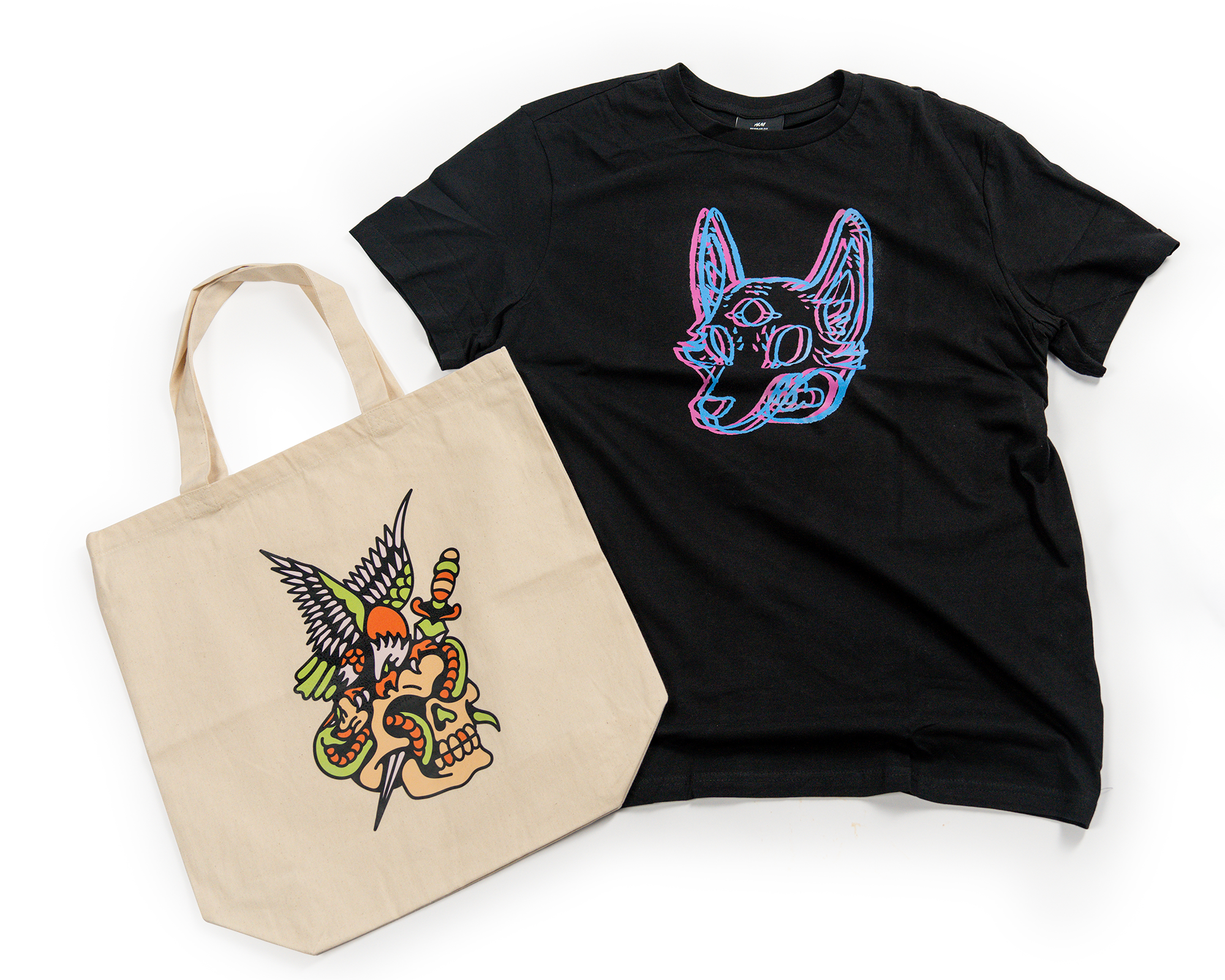 Black t-shirt with neon animal face design and beige tote bag with colorful skull, wings, and snake illustration on white background.