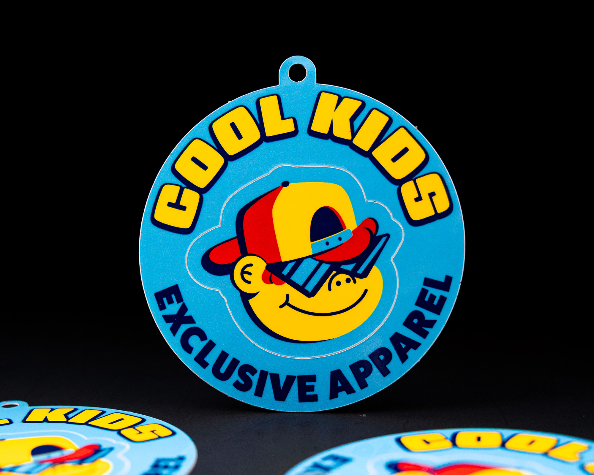 A blue and yellow circular hang tag reads "Cool Kids Exclusive Apparel" with a cartoon character wearing a cap and sunglasses.