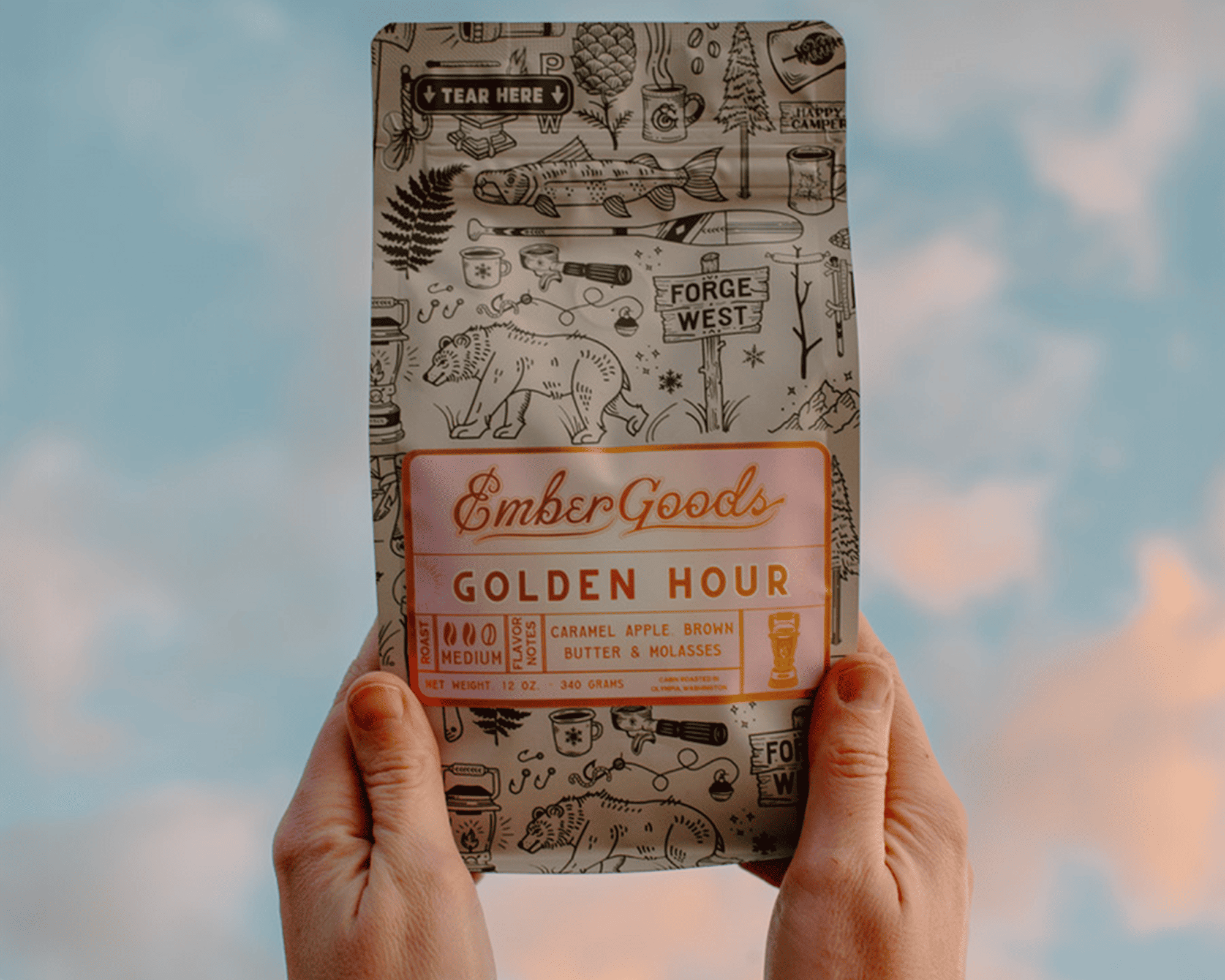 Mani che reggono una busta di caffè Golden Hour di Ember Goods con etichetta metallizzata su uno sfondo di cielo