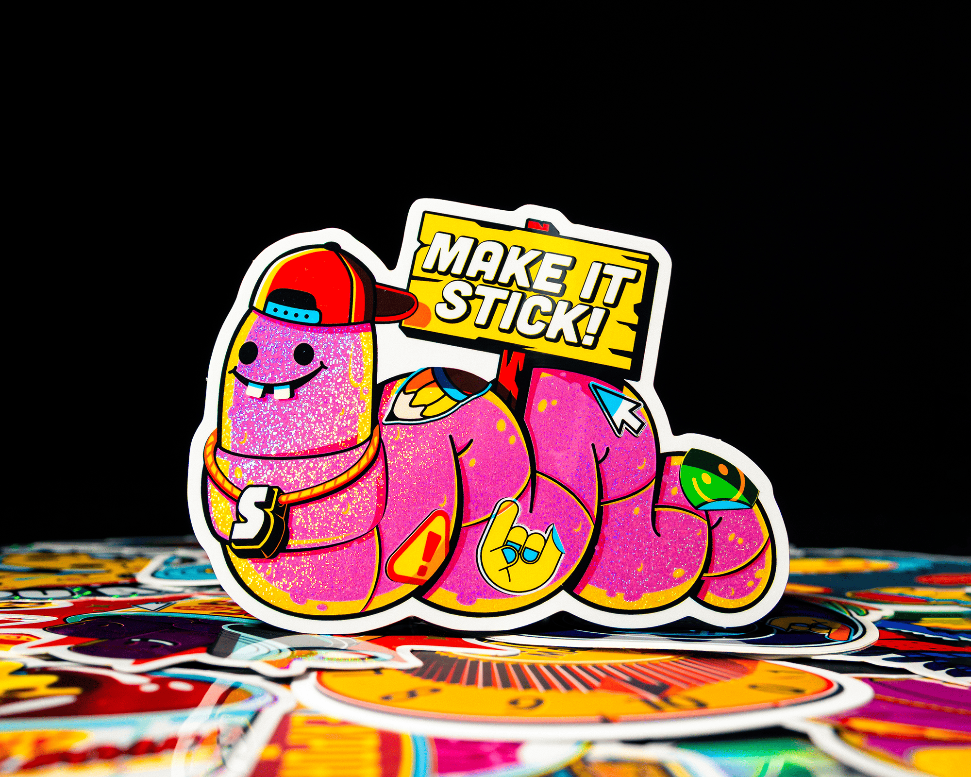 A colorful sticker that is a worm holding a sign that say "MAKE IT STICK!"
