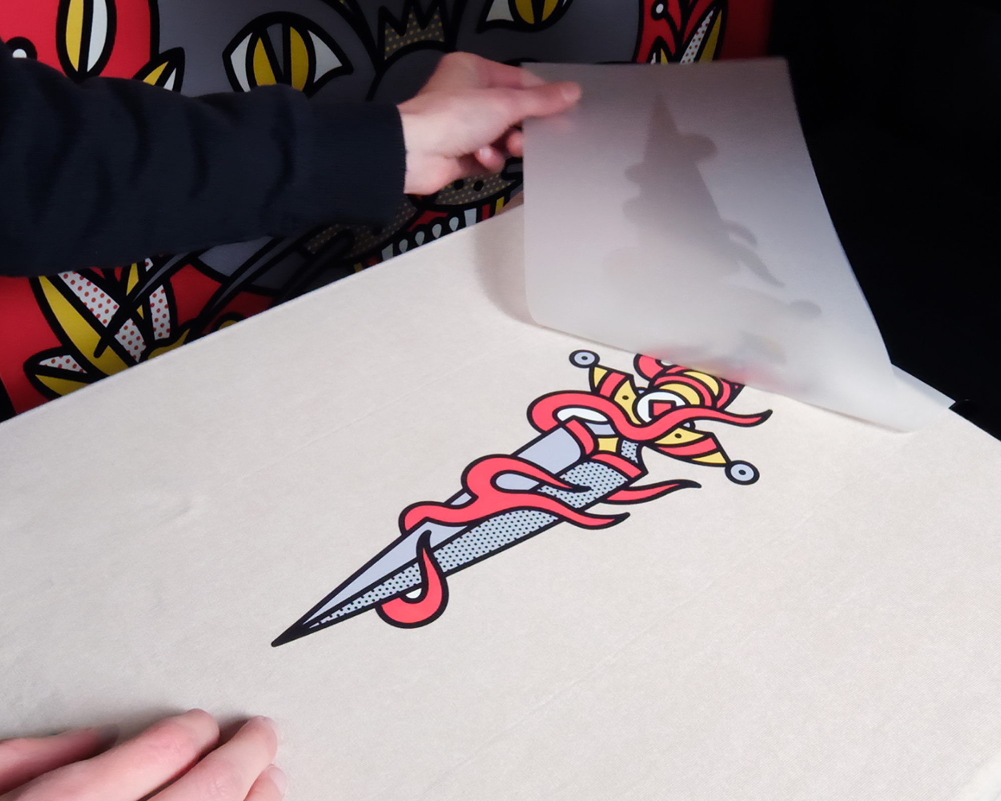 Hands peeling transfer paper off a fabric, revealing a colorful design of a dagger entwined with a snake, against a vibrant background.