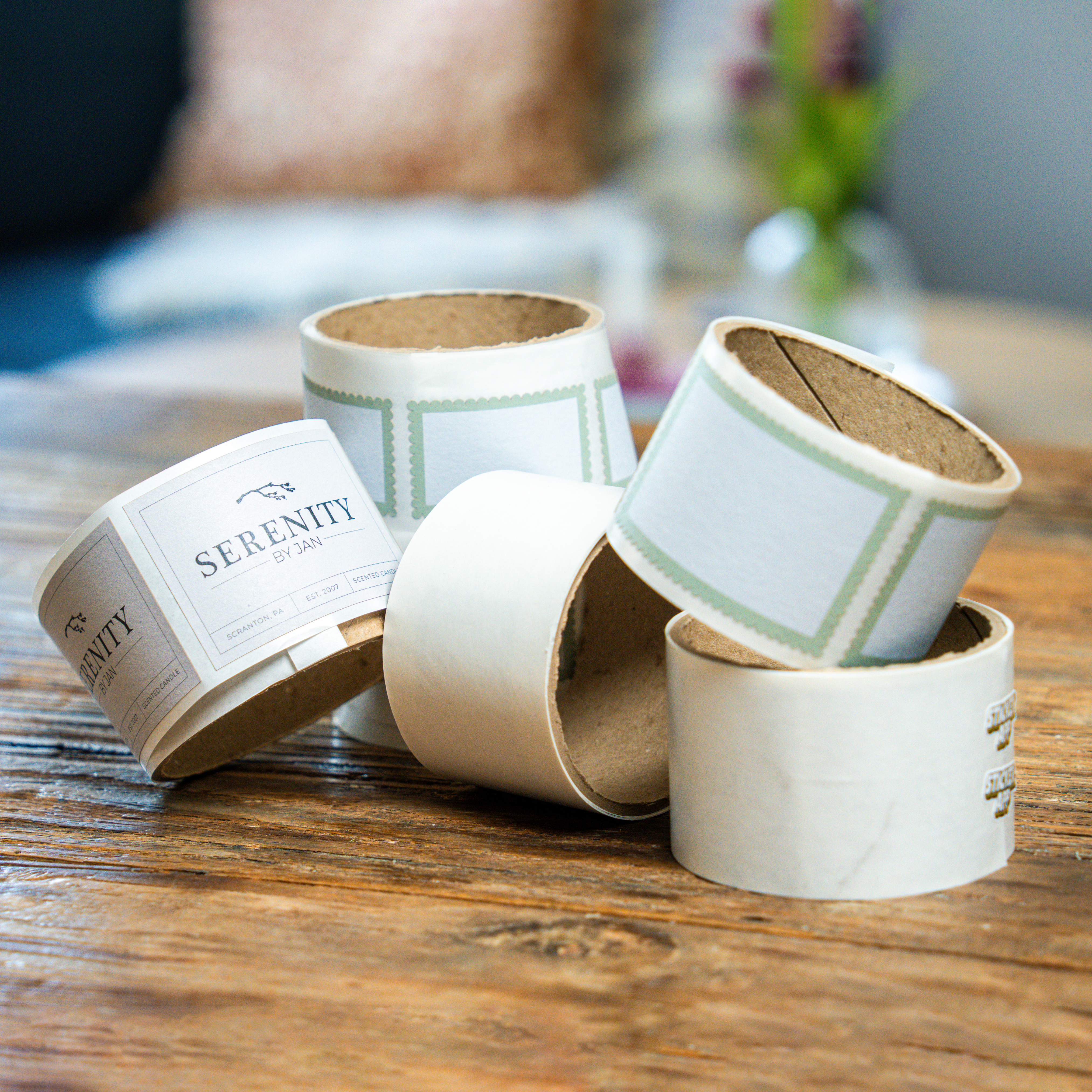 Rolls of printed labels scattered on a wooden table, including minimalist product labels and blank bordered sticker designs.