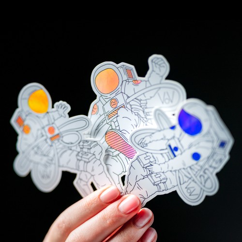 A hand holding a holographic sticker with three cartoon space mans on it